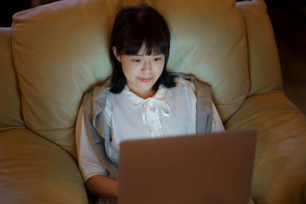 照明の低い部屋でソファの上にノートパソコンを使っている若い女性 - 写真・画像