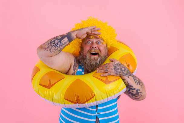 El hombre gordo sorprendido con la peluca en la cabeza está listo para nadar con un salvavidas de rosquillas - Foto, imagen