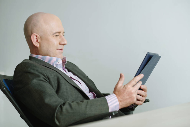 Зміст розслабленого лисого бізнесмена в куртці, що сидить в офісному кріслі і використовує планшет під час роботи в Інтернеті в офісі
 - Фото, зображення