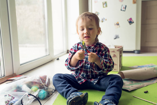 Kahden vuoden ikäinen poika istuu lattialla erilaisten ovelien materiaalien joukossa ja pitelee pihdeillä varustettua nallekarkkia. Luova lapsuus. - Valokuva, kuva