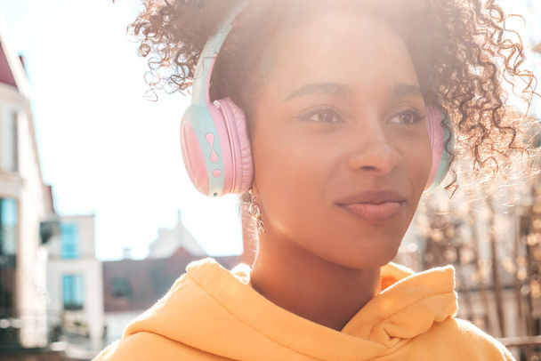 Beautiful black woman with afro curls hairstyle.Smiling model in yellow hoodie.Sexy carefree female enjoying listening music in wireless headphones.Posing on street background at sunset - Фото, изображение