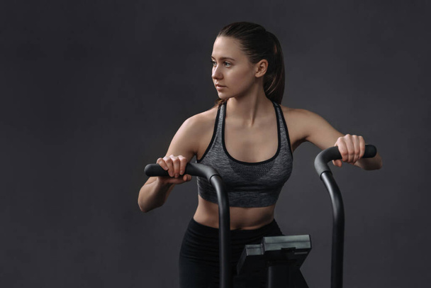 Attractive young woman in sportswear using exercise bike for cardio workout at fitness gym. Fit pretty female having air bike class. Copy space, isolated on gray wall background - 写真・画像
