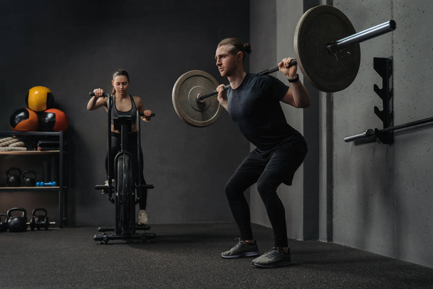 Group crossfit training. Muscular man and beautiful woman working out with crossfit equipment in gym. Young couple have functional training. Guy is lifting barbell while girl warming up on air bike - Photo, Image