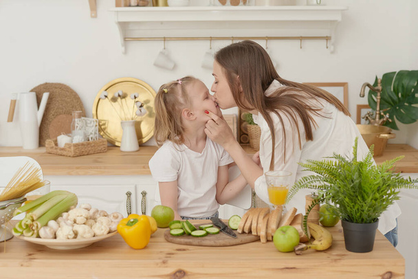 Mère joyeuse et petite fille préparent la salade ensemble dans la cuisine et s'amusent. La fille embrasse sa mère à la maison - Photo, image