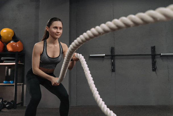 Portret van een fitnessvrouw met zware touwen die traint in de functionele trainingscrossfit dark gym. Atletische jonge vrouw doet workout in functionele trainingsfitnessruimte met behulp van battle touwen - Foto, afbeelding