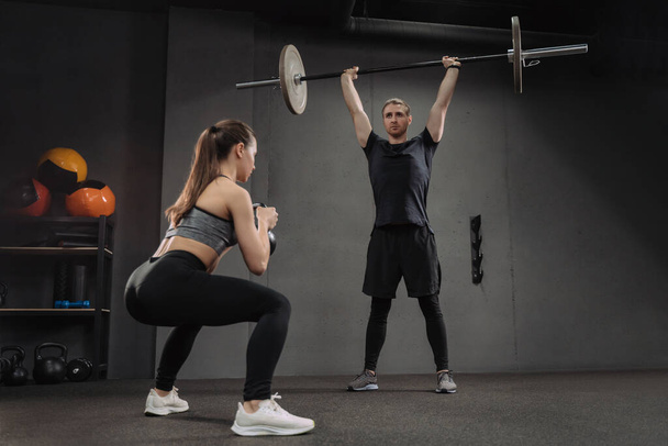 Fit caucasian couple doing crossfit exercises at dark gym. Attractive sporty woman doing squats with kettlebelll while muscular man practicing with heavy barbell. Man and woman having workout together - Photo, Image