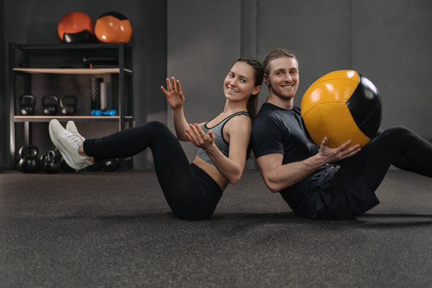 Happy crossfit ζευγάρι κατάρτισης μαζί στο σκοτεινό γυμναστήριο με επαγγελματικό αθλητικό εξοπλισμό στο παρασκήνιο. Fit άνθρωπος και σπορ γυναίκα κάνει κοιλιακούς άσκηση μαζί χρησιμοποιώντας ιατρική μπάλα, δίνοντας ο ένας τον άλλον - Φωτογραφία, εικόνα