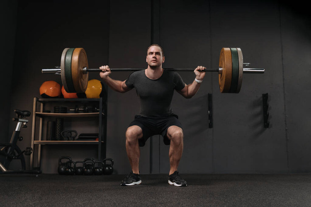 Muscular crossfit athlete doing weight lift squats with barbell at dark gym. Man lifting a weight in the sport club, have a functional training class, working out - Photo, Image