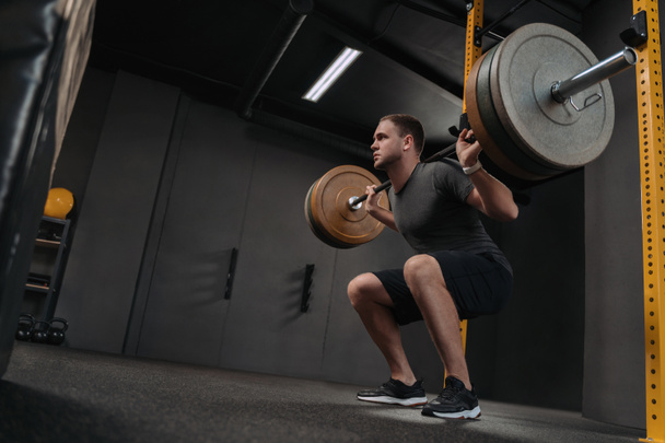 Homme caucasien musclé soulevant des poids et faisant squat dos dans la salle de gym. Crossfit athlète tient un haltère lourd sur l'épaule derrière le cou tout en s'accroupissant. Fitness et entraînement - Photo, image