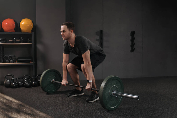 Crossfit athlete squatting and preparing to lift heavy barbell while training weightlifting. Fit young man lifting weight, working out at gym. Dark background. Sport, fitness, powerlifting concept - Photo, Image
