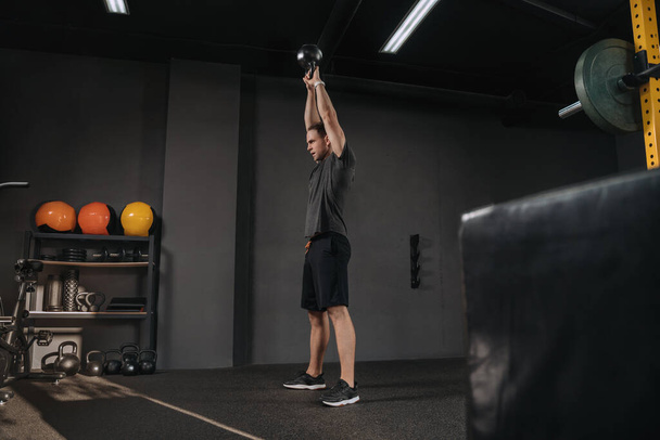 Handsome athletic young man lifting kettlebell with two hands as crossfit workout. Muscular man holding kettlebell overhead at gym. - Photo, Image