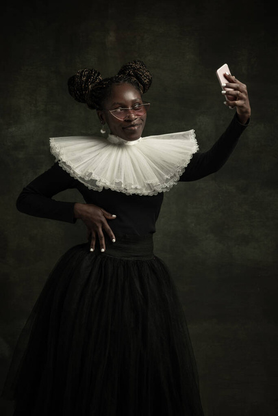 Portrait of medieval African young woman in black vintage dress with big white collar posing isolated on dark green background. Taking selfie - 写真・画像