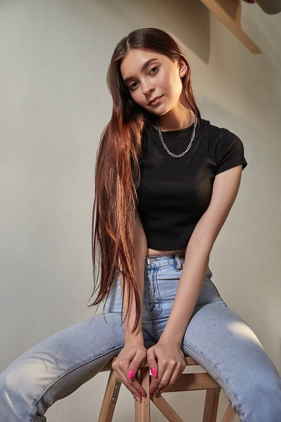 portrait of young caucasian girl with long hair in jeans sitting on wooden stool - Photo, Image