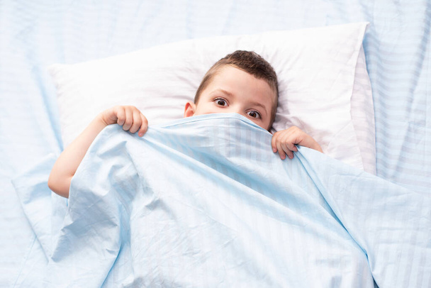 Ein verängstigter vierjähriger Kleinkind-Junge verdeckte sein Gesicht mit einer blauen Decke. Blick unter die Abdeckungen. - Foto, Bild