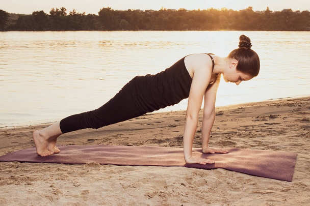 A woman stands in a plank pose on the river bank on the beach at sunset in summer. Yoga or fitness classes in nature. - Photo, Image