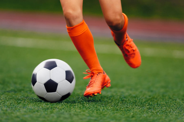 Ποδόσφαιρο παίκτης χτυπά την μπάλα στο τεχνητό χλοοτάπητα. Ποδοσφαιριστής με αθλητικά παπούτσια κλωτσάει μπάλα. Πόδια του ποδοσφαιριστή που παίζουν αγώνα ανταγωνισμού - Φωτογραφία, εικόνα