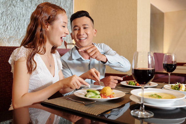 Happy smiling young man feeding girlfriend with piece of meat from his plate - Photo, Image