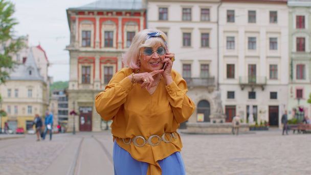 都会の通りを歩きながら携帯電話で話すスタイリッシュな服のシニア観光客の女性 - 写真・画像