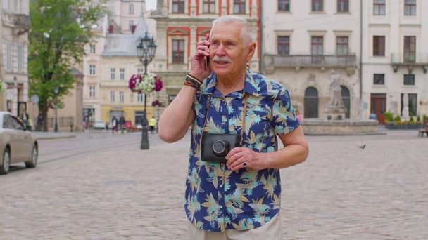 Старший старый турист в стильной одежде разговаривает по мобильному телефону во время прогулки по городской улице - Фото, изображение