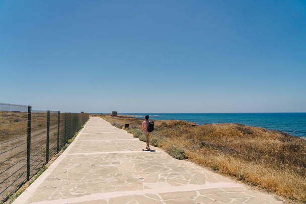 バックパック付きの男の観光客は、夏の晴れた暑い日にキプロスのパフォスで地中海の岩場の海岸を歩く道に沿って歩く。海岸沿いの歩道。キプロス島を徒歩で探索してください。ハイキング - 写真・画像