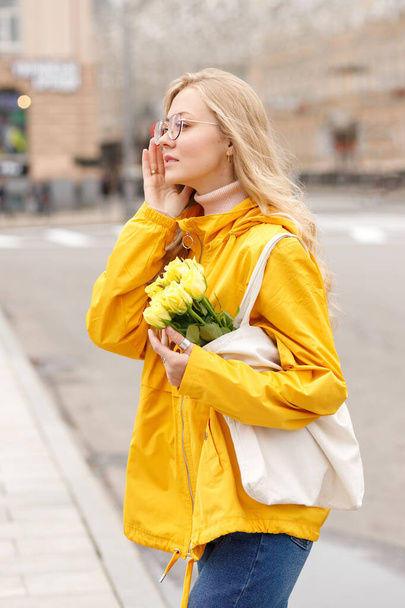 Девушка в желтой куртке с цветами в эко-сумке в городе осенью - Фото, изображение