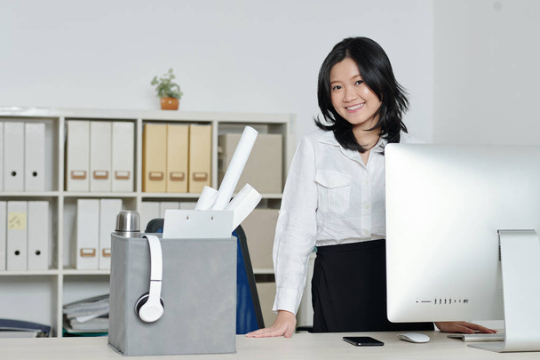 Glücklich schöne junge Frau steht an ihrem neuen Schreibtisch mit Computer und Kiste voller Sachen - Foto, Bild