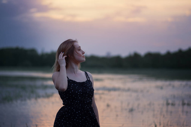 Summer Portrait of a beautiful young woman in a black dress . Standing in the water against the backdrop of a summer sunset at dusk - Photo, Image