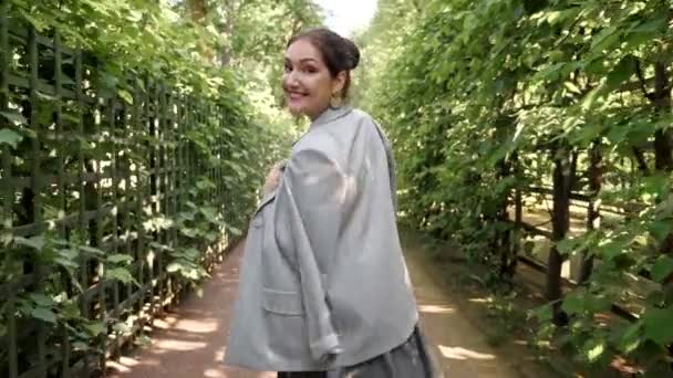 Νεαρή μελαχρινή γυναίκα περπατά στον κήπο και γυρίζει, χαμογελώντας - Πλάνα, βίντεο