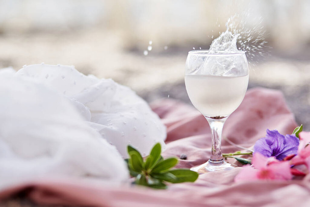 Explosion von Tonic Cocktail im Weinglas am Strand mit Blumen. Sommercocktail. Zarte Seidenmaterialien und Bokeh-Hintergrund. Erfrischendes Sommerkonzept. Hochwertiges Foto - Foto, Bild