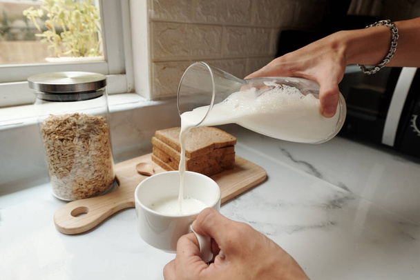 крупным планом неузнаваемого человека, стоящего у прилавка и наливающего молоко в чашку во время приготовления завтрака - Фото, изображение