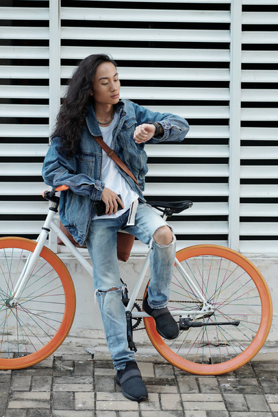 Hombre vietnamita joven guapo serio con el pelo ondulado sentado en el estante de la bicicleta y el mensaje de lectura en smartwatch en la ciudad - Foto, Imagen