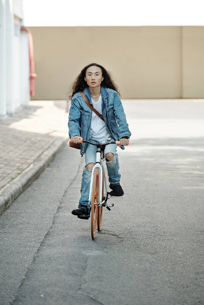 Aktif, genç Vietnamlı uzun saçlı, dizlerinde delikler olan kot giyen şehirde bisiklet sürerken etrafa bakan adam. - Fotoğraf, Görsel