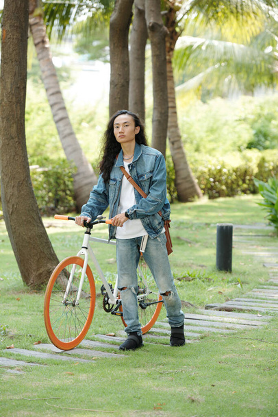 Serious young Vietnamese guy in denim wear walking with bicycle on pathway in tropical park with palms - Photo, Image