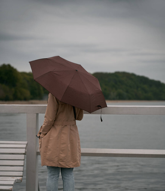 Back of lonely woman with umbrella on the wooden pier looking distant to the seashore. Rainy weather and loneliness concept. Raining, sea and long haired brunette with umbrella. - Fotoğraf, Görsel