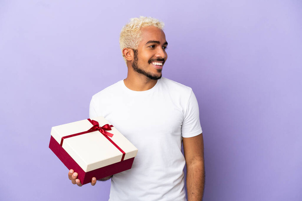Nuori kolumbialainen mies, jolla on lahja, joka on eristetty violettiin taustaan katsoen sivulle ja hymyillen - Valokuva, kuva