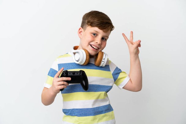 Kleine roodharige jongen met een game pad geïsoleerd op een witte achtergrond glimlachen en tonen overwinning teken - Foto, afbeelding