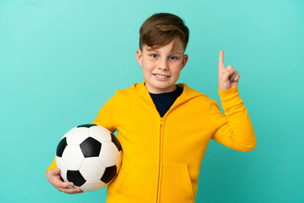 Рыжий ребенок играет в футбол изолированы на синем фоне указывая на отличную идею - Фото, изображение
