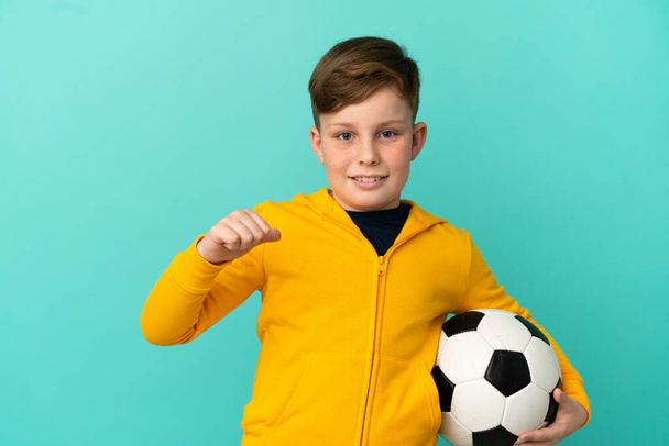 Mavi arka planda izole bir şekilde futbol oynayan kızıl saçlı çocuk gururlu ve kendini beğenmiş. - Fotoğraf, Görsel