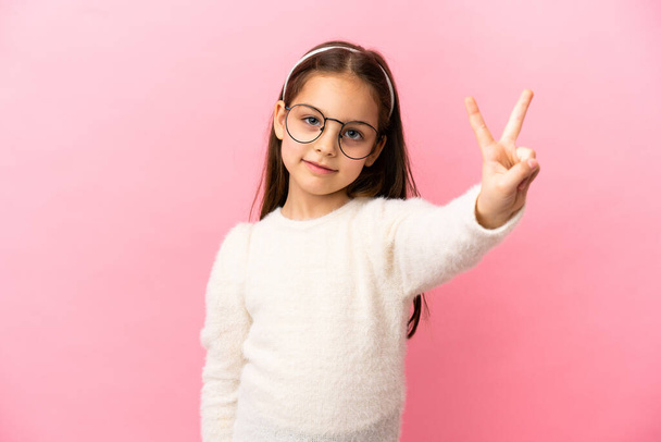 Маленька біла дівчинка ізольована на рожевому фоні посміхається і показує знак перемоги
 - Фото, зображення