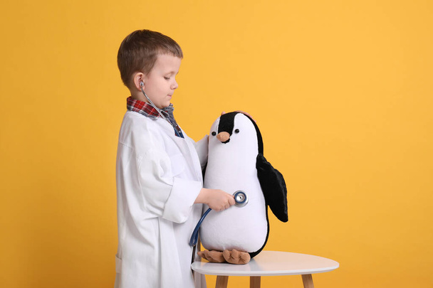 Cute little boy in pediatrician's uniform playing with stethoscope and toy penguin on yellow background - Photo, Image