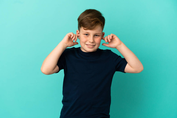 Little redhead boy isolated on blue background frustrated and covering ears - Φωτογραφία, εικόνα