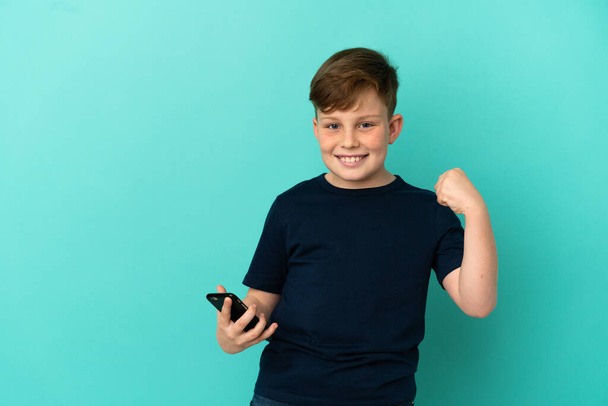 Piccolo ragazzo rosso isolato su sfondo blu con telefono in posizione di vittoria - Foto, immagini