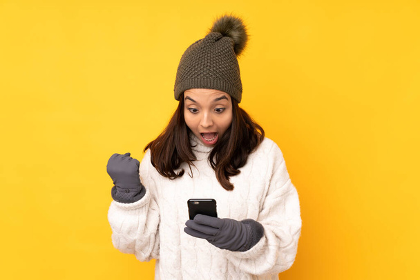 Молодая женщина в зимней шляпе на изолированном желтом фоне удивлена и отправить сообщение - Фото, изображение