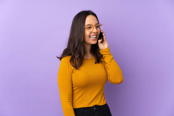 Молодая женщина смешанной расы изолированы на фиолетовом фоне ведения разговора с мобильным телефоном - Фото, изображение