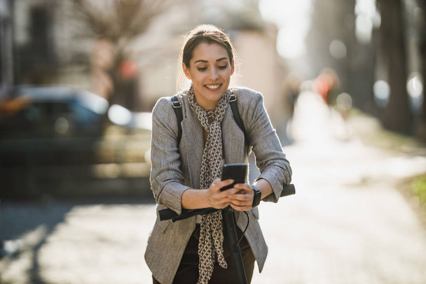 Eine junge Geschäftsfrau mit Smartphone auf dem Weg zur Arbeit mit einem Elektroroller. - Foto, Bild