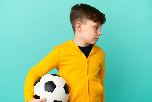 Kleiner rothaariger Junge isoliert auf blauem Hintergrund mit Fußball - Foto, Bild