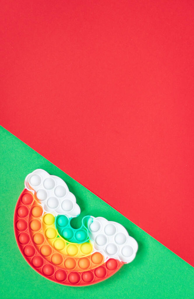 Veelkleurige siliconen speelgoed antistress pop het in de vorm van een regenboog met wolken op een groene rode diagonale achtergrond - Foto, afbeelding