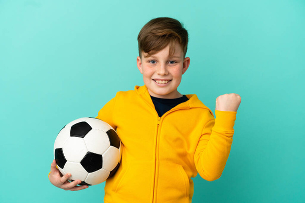 Redhead dítě hraje fotbal izolované na modrém pozadí slaví vítězství v pozici vítěze - Fotografie, Obrázek
