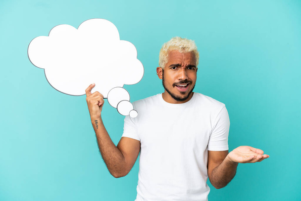 Young Colombian handsome man isolated on blue background holding a thinking speech bubble and with sad expression - 写真・画像
