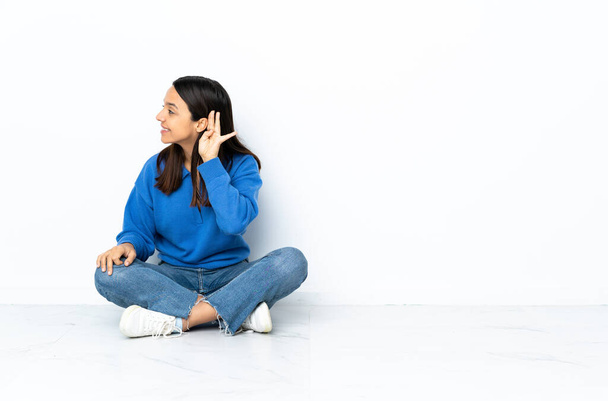 Nuori sekarotuinen nainen istuu lattialla eristyksissä valkoisella taustalla kuunnellen jotain laittamalla käden korvaan. - Valokuva, kuva